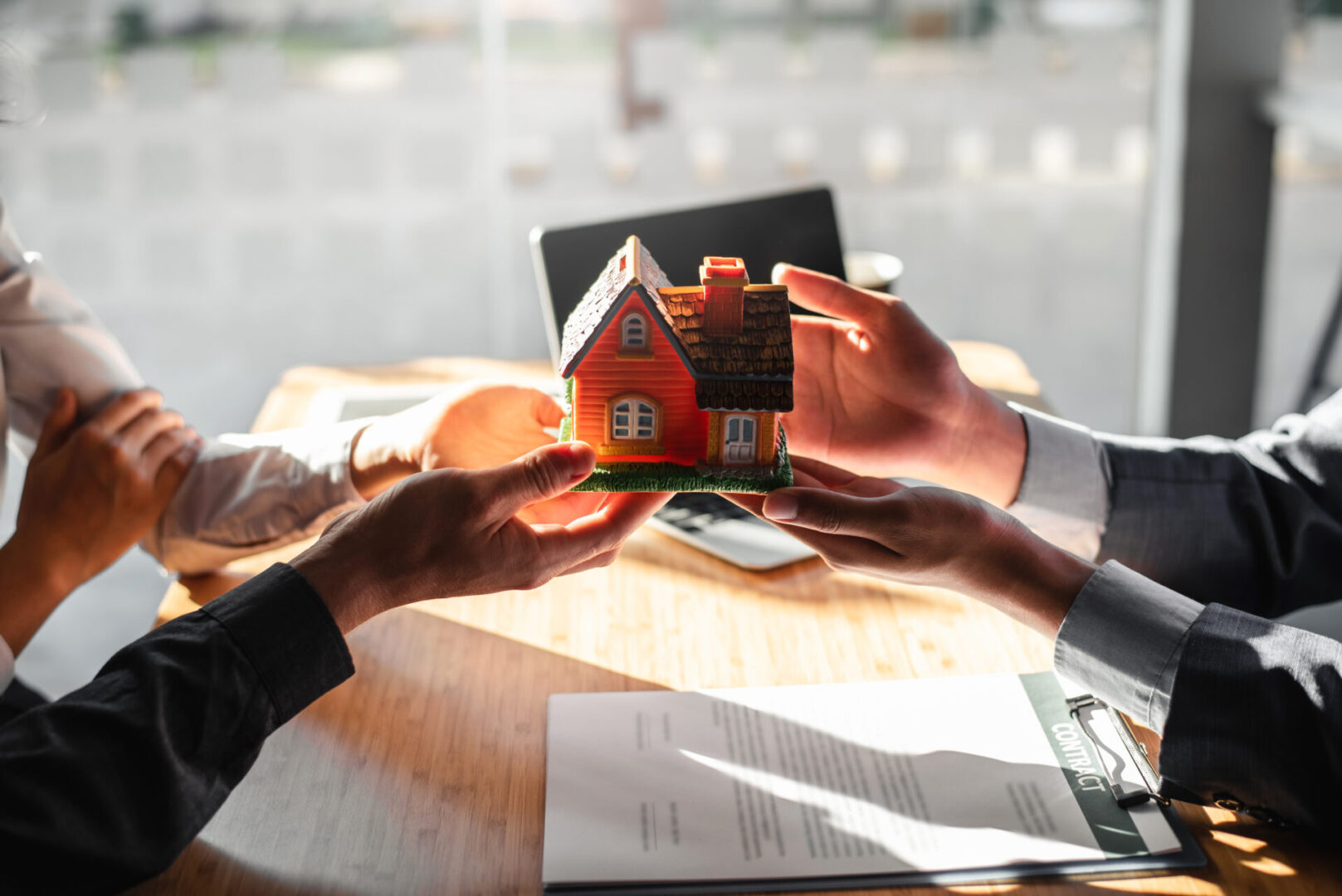 Two people exchanging a miniature house model over a table with paperwork.